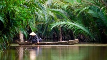 A person in a traditional conical hat paddles a wooden boat through a lush, green river (Photo: Nikhita Singhal via Unsplash)